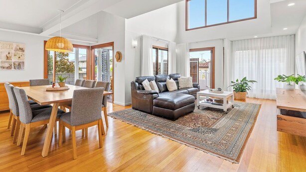 A spacious living room with a wooden dining table, grey upholstered chairs, a brown leather sofa, and a large window providing natural light. - Beach House - Lennox Head (Lennox head)