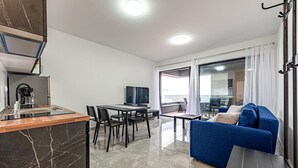 Un soggiorno moderno con un divano blu, un'area pranzo con tavolo e sedie, una cucina con macchina del caffè e un'ampia finestra che lascia entrare luce naturale.