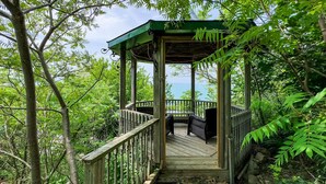 A wooden gazebo with a covered deck, surrounded by lush greenery and overlooking a body of water.
