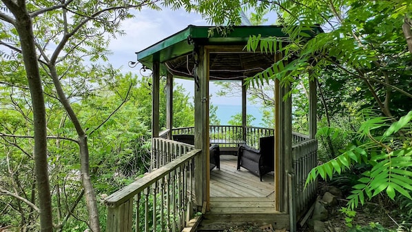 A wooden gazebo with a covered deck, surrounded by lush greenery and overlooking a body of water.