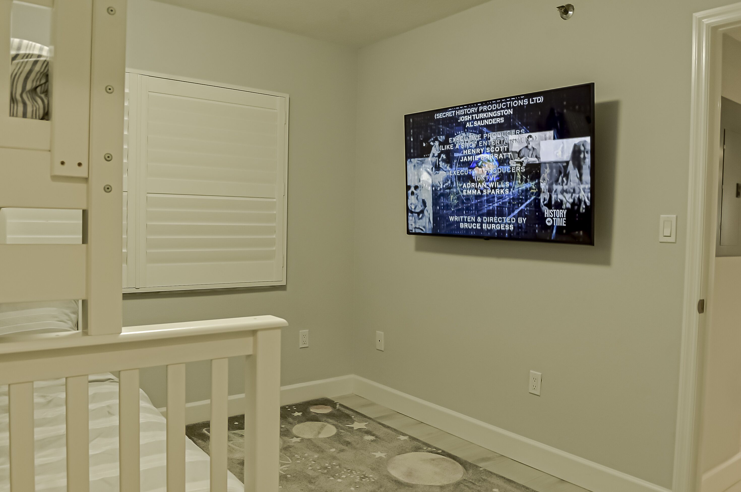Un dormitorio con una cuna, un televisor instalado en la pared mostrando una película y una ventana con persianas.