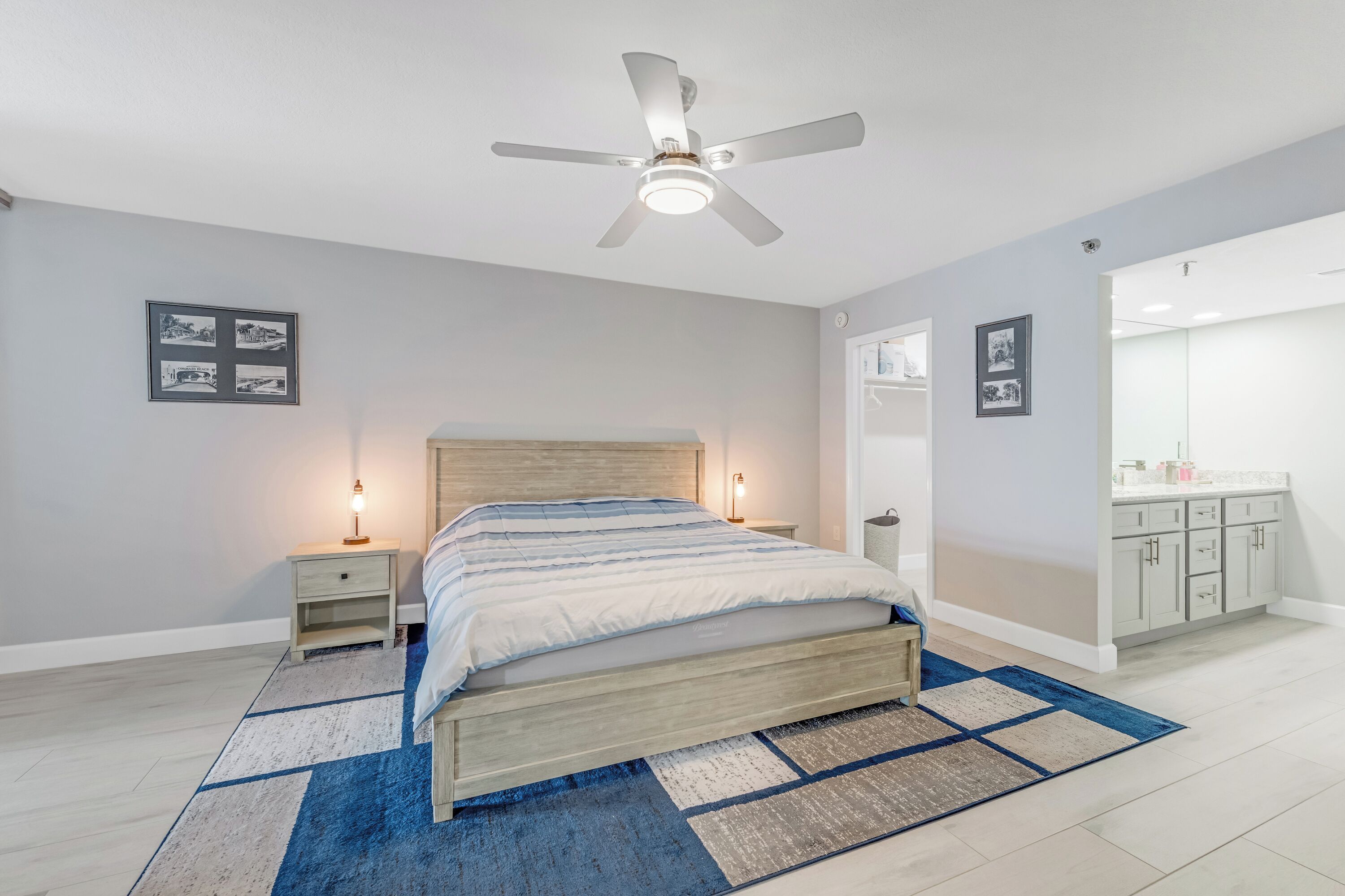 A bedroom with a bed, nightstands, a ceiling fan, a rug, and a bathroom with a mirror and cabinets.