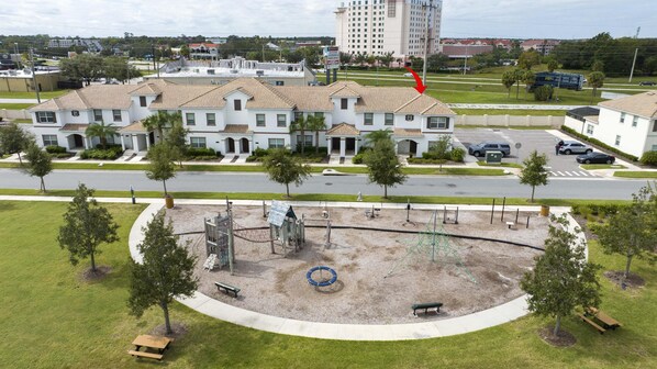 Zona residencial con una casa grande de color blanco y techo de tejas, un parque infantil con columpios y tobogán, y un estacionamiento con coches.