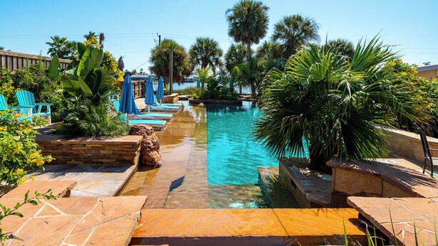 A pool area with clear water, blue lounge chairs, and palm trees. - Little Bay Club by Miss Kitty’s (Rockport)