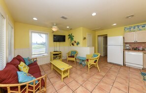 A compact living space with a sofa, two chairs, a small table, a ceiling fan, a TV, and a kitchen area with a fridge and a coffee maker. - Little Bay Club by Miss Kitty’s (Rockport)