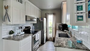 A modern kitchen with white cabinetry, stainless steel appliances, granite countertops, and a view of the outdoors through a window. - Rockport Racquet and Yacht Club by Miss Kitty’s (Rockport)