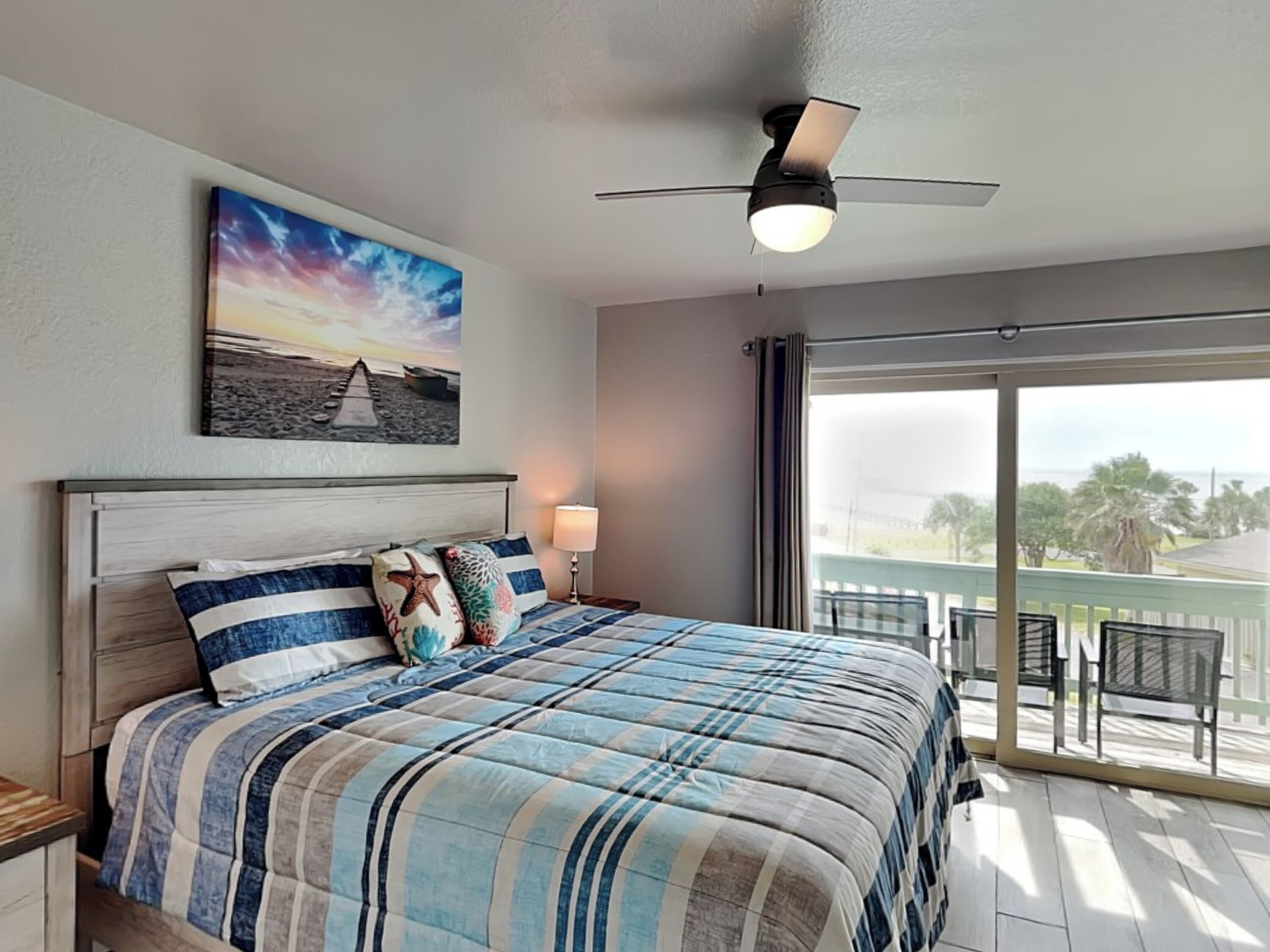 Quarto com uma cama grande, um ventilador de teto, uma pintura de uma cena de praia e vista para o exterior através de uma porta de correr de vidro.