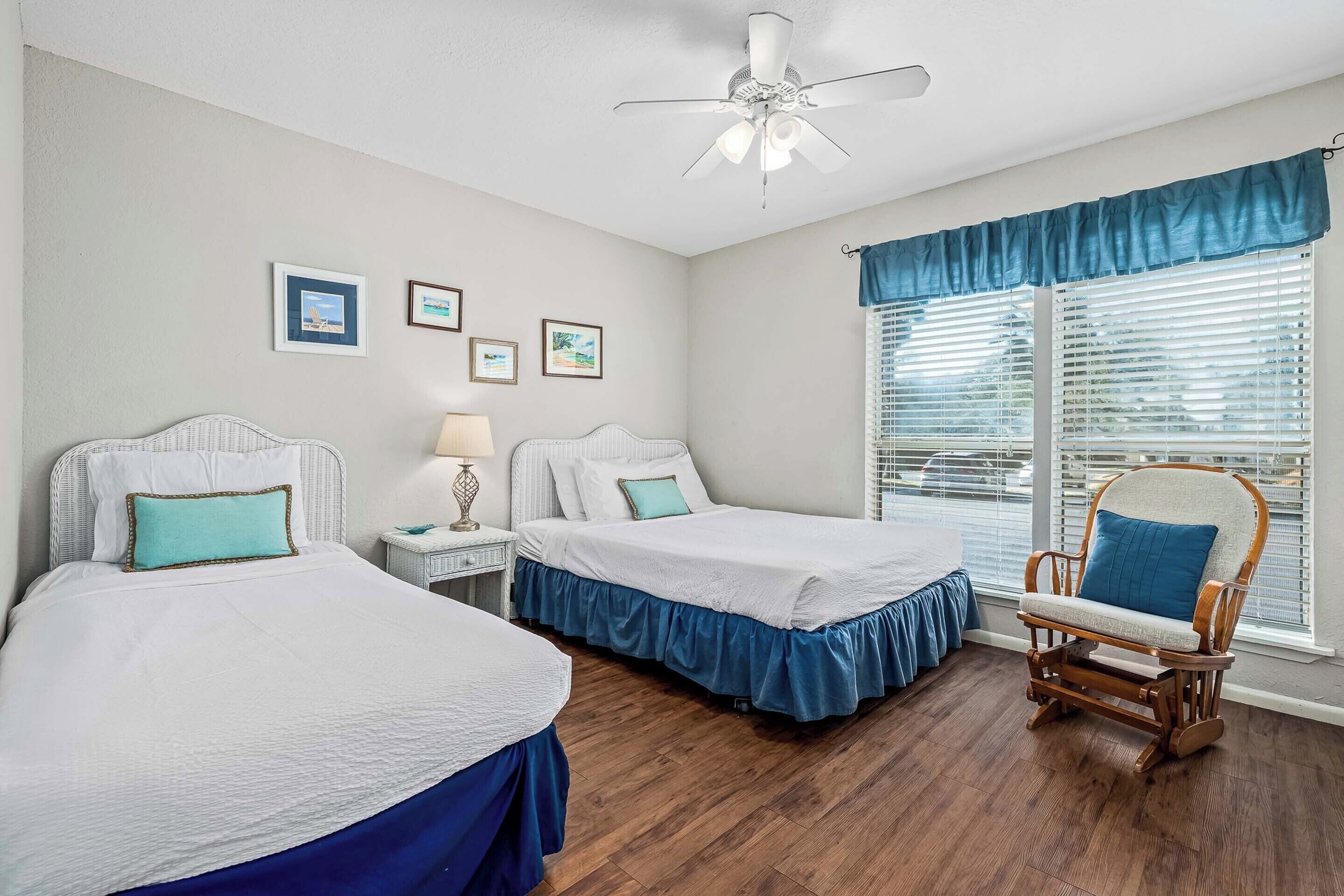 A bedroom with two beds, a rocking chair, a ceiling fan, and a window with blinds.
