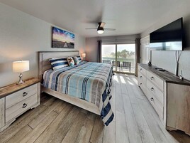 Quarto com cama, mesa de cabeceira, cômoda, televisão e varanda com vista.
