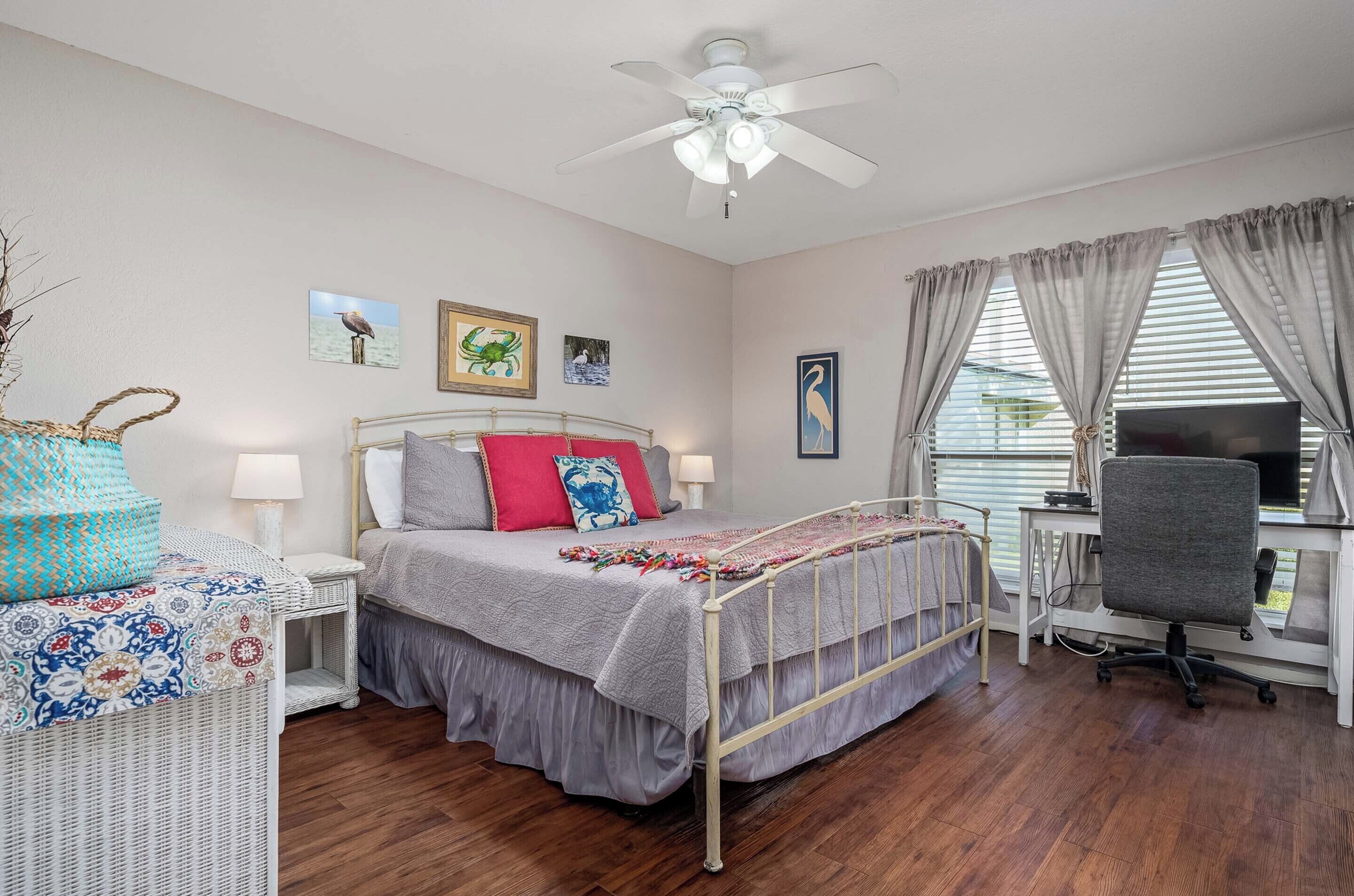 A bedroom with a bed, bedside tables, a desk, a chair, a ceiling fan, and artwork on the walls.