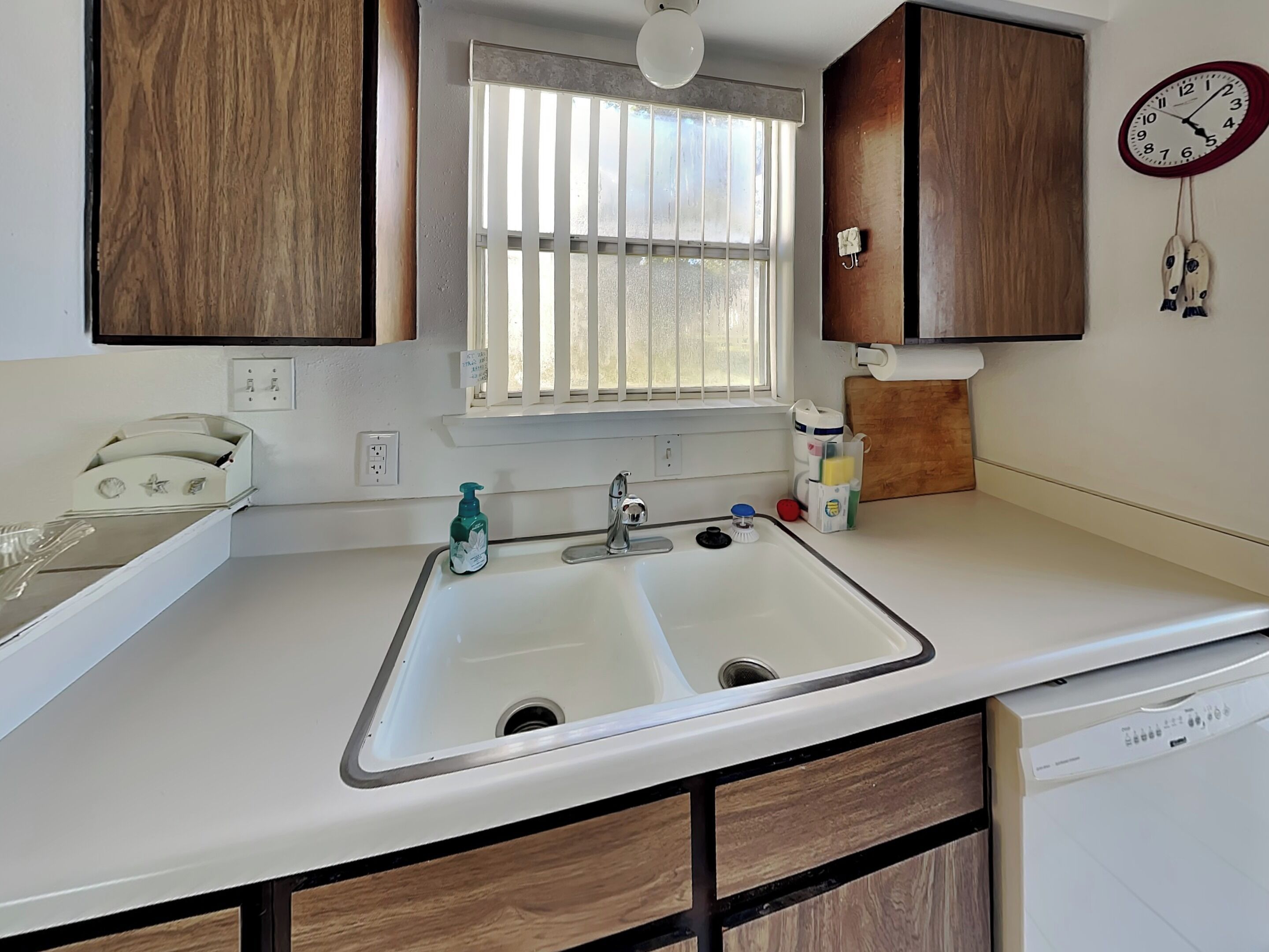 A kitchen with a double sink, wooden cabinets, a window with blinds, a clock, and a dishwasher.