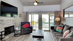 A living room with a fireplace, a sofa, a coffee table, a TV mounted on the wall, and a ceiling fan. - Rockport Racquet and Yacht Club by Miss Kitty’s (Rockport)