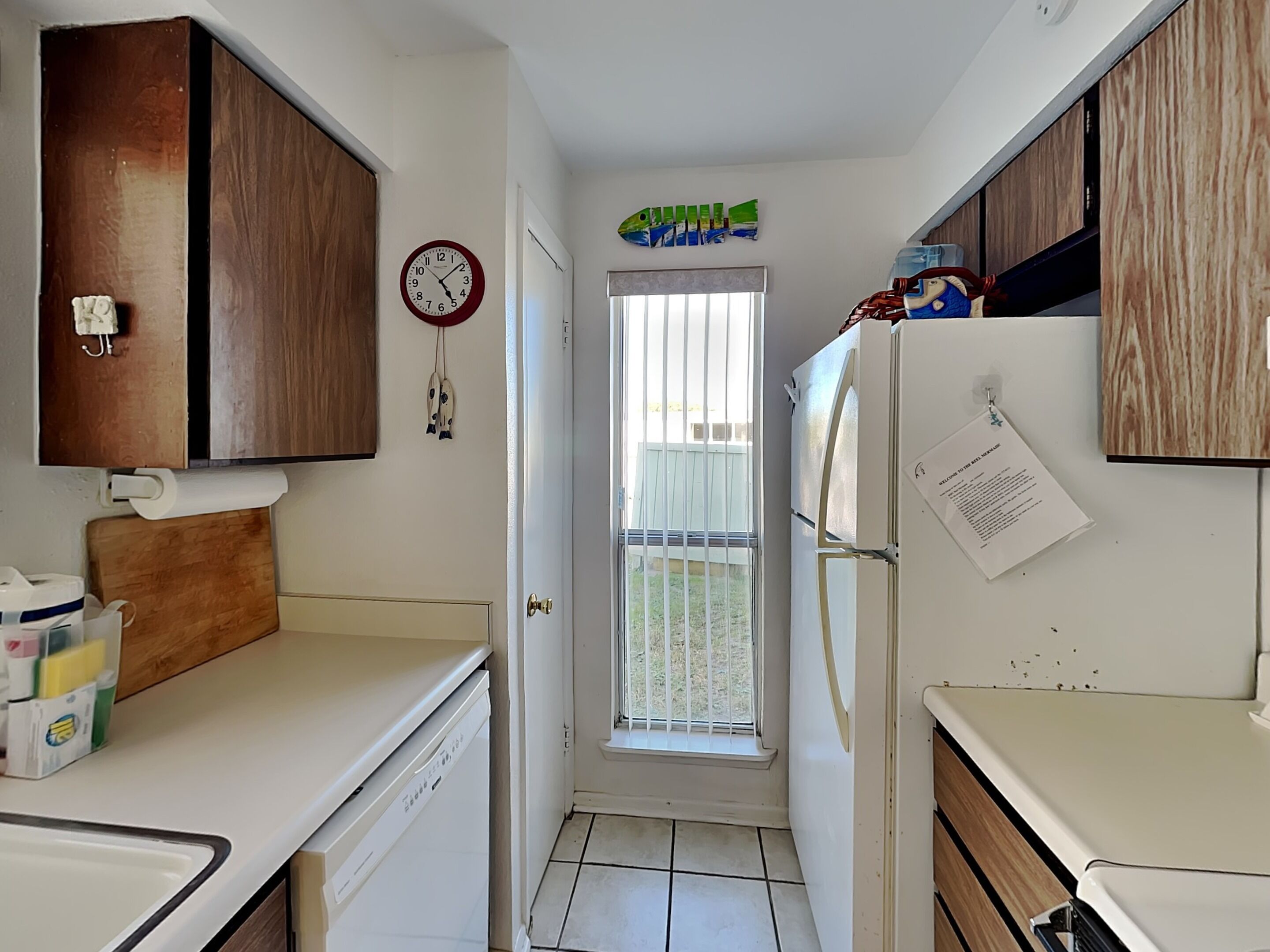 A compact kitchen with wooden cabinets, a white countertop, a clock, and a door leading to a balcony.