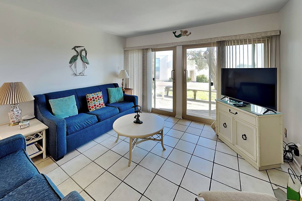 A living room with a blue sofa, a white coffee table, a TV on a cabinet, and a sliding glass door leading to an outdoor area.