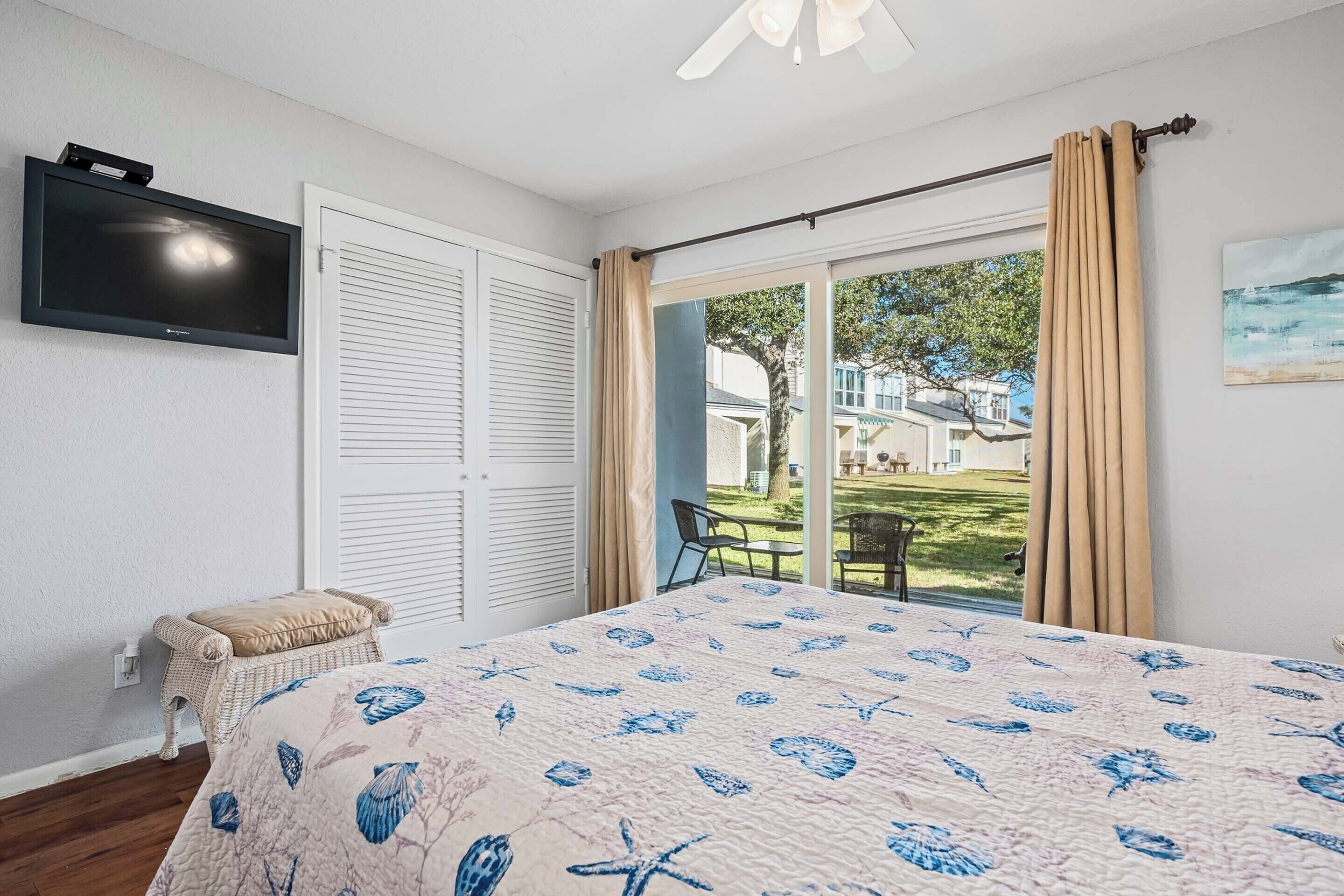 A bedroom with a bed, a television, a window with curtains, and a view of the outdoors.