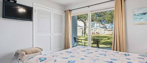 A bedroom with a bed, a television, a window with curtains, and a view of the outdoors.