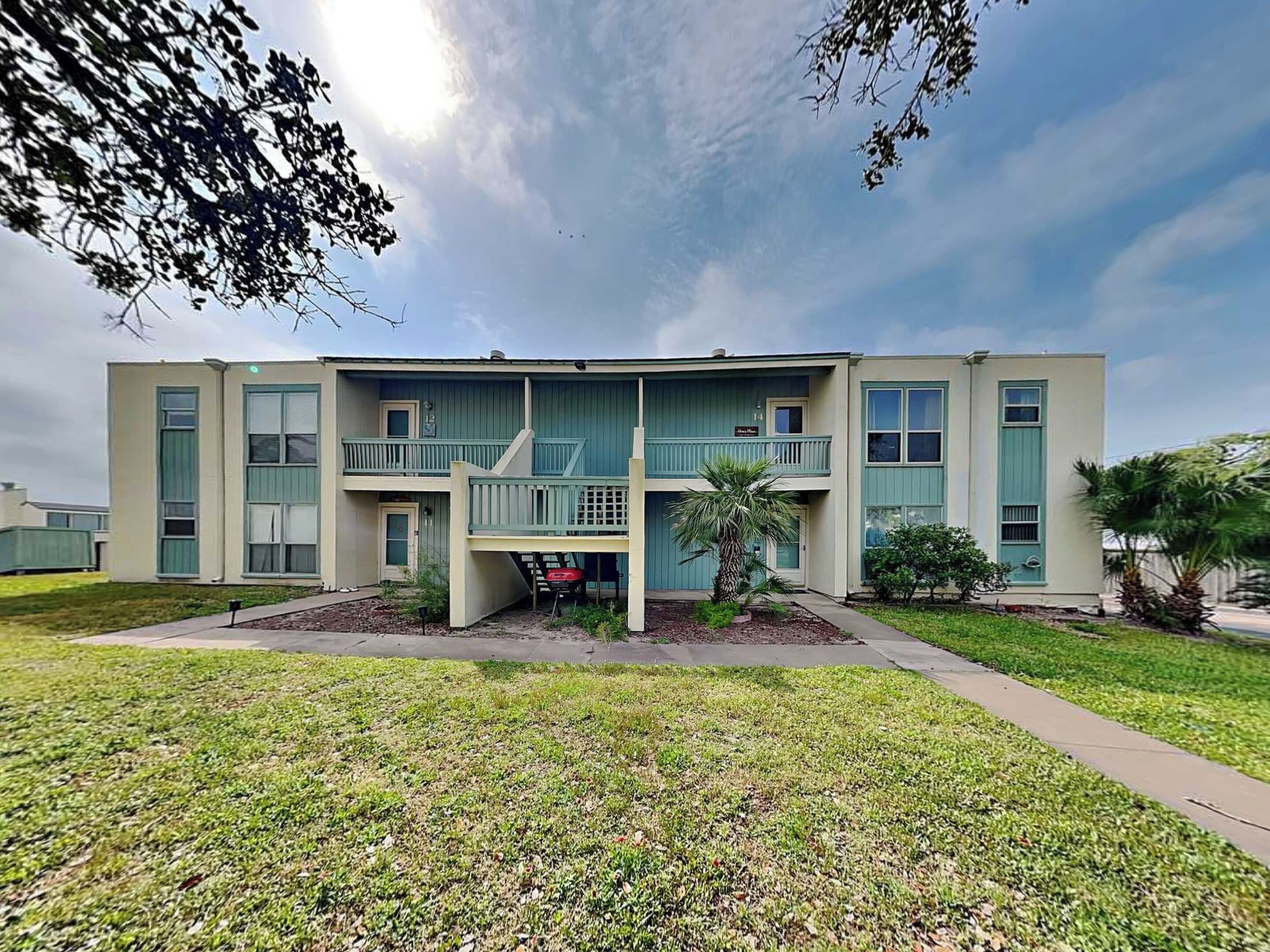A two-story building with a flat roof, a central entrance, and a small garden area with grass and a palm tree.