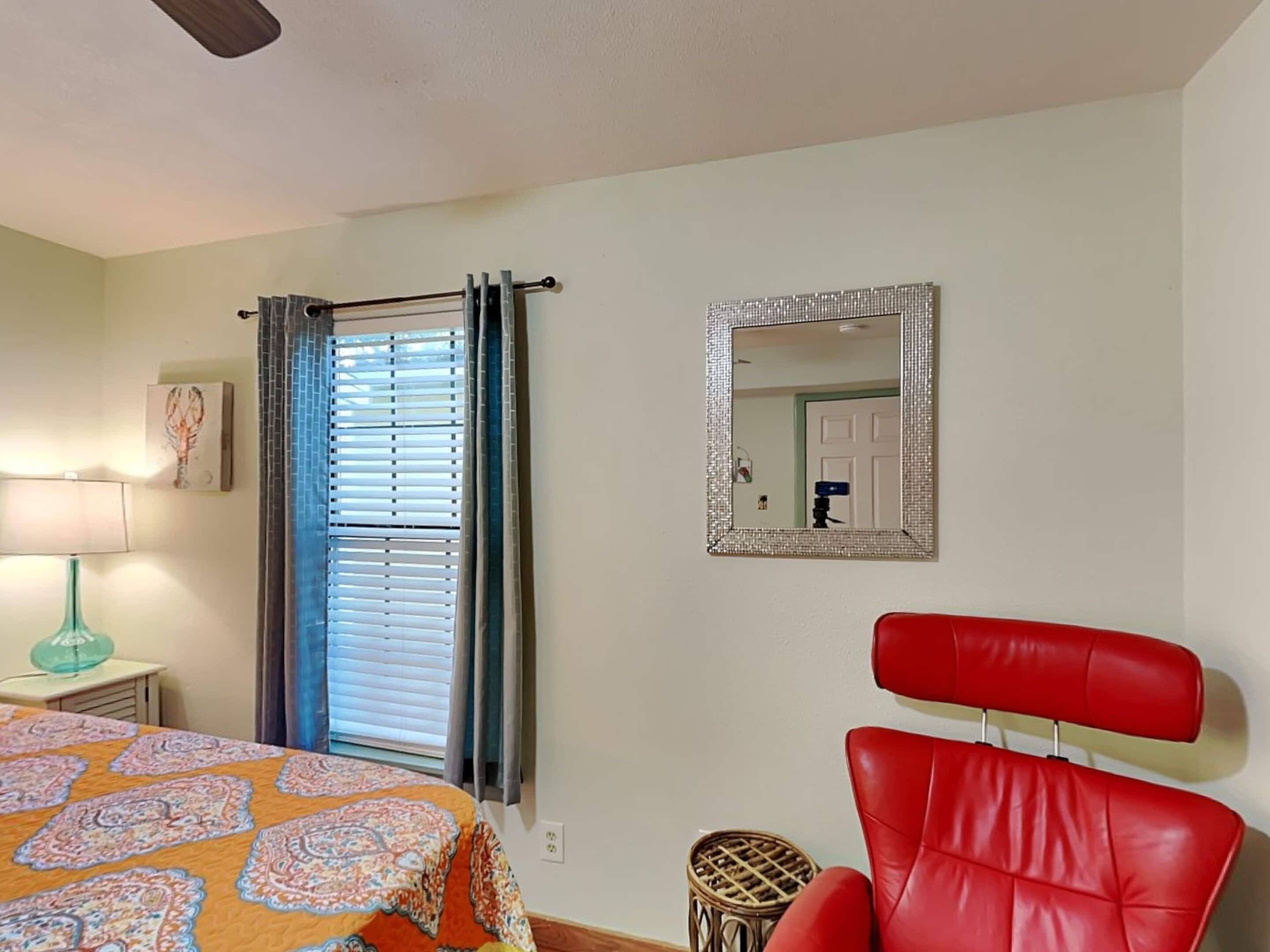 Un dormitorio con cama, una silla roja, un espejo y una ventana con persianas.