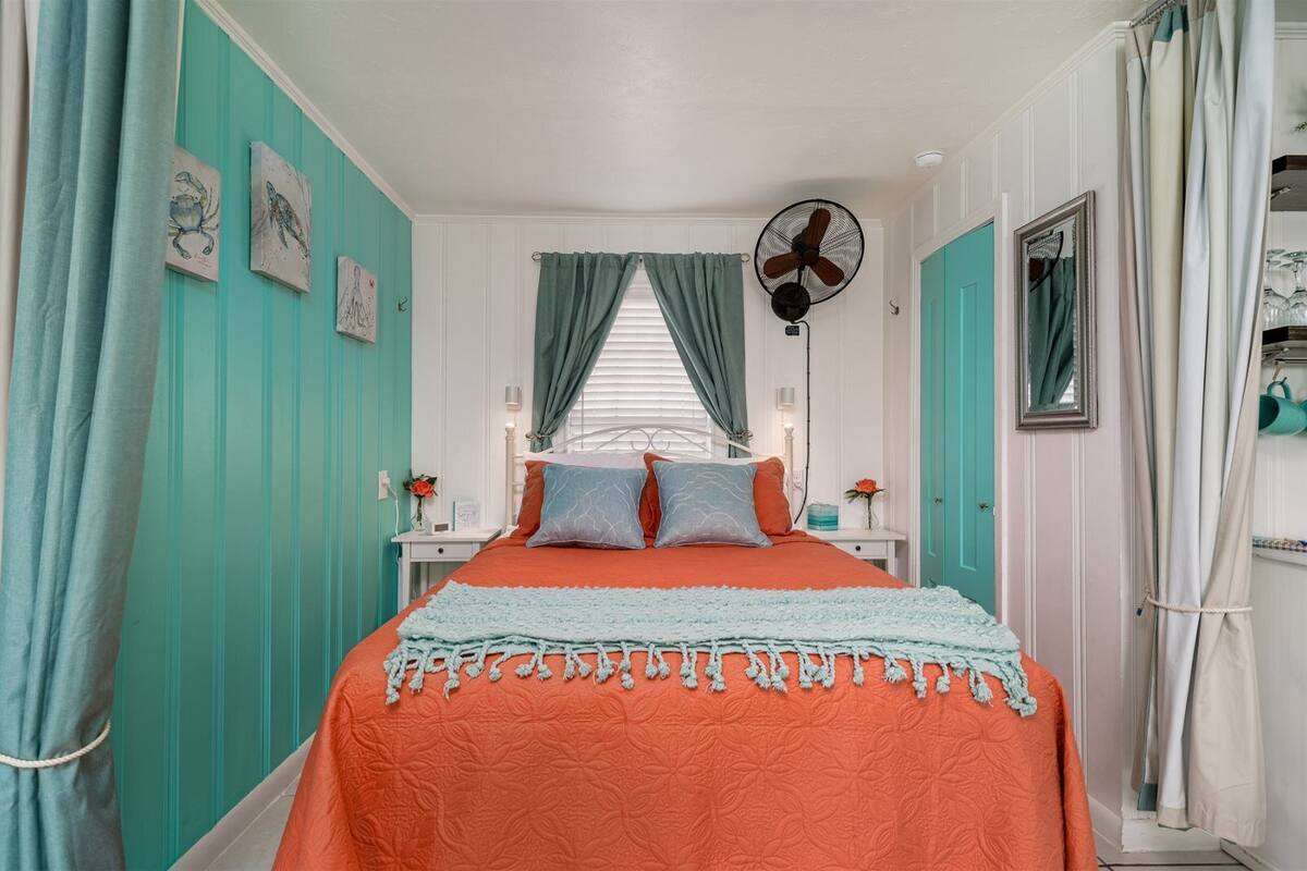 Un dormitorio con paredes verde azulado, una cama con ropa de cama naranja, un ventilador de techo y una ventana con cortinas.