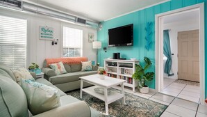 A living room with a teal accent wall, a sofa, a coffee table, a TV, and a bookshelf. - Sun Harbor Cottages by Miss Kitty’s (Rockport)