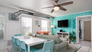 A living room with teal walls, a ceiling fan, a TV, and a dining area with a white table and teal chairs. - Sun Harbor Cottages by Miss Kitty’s (Rockport)
