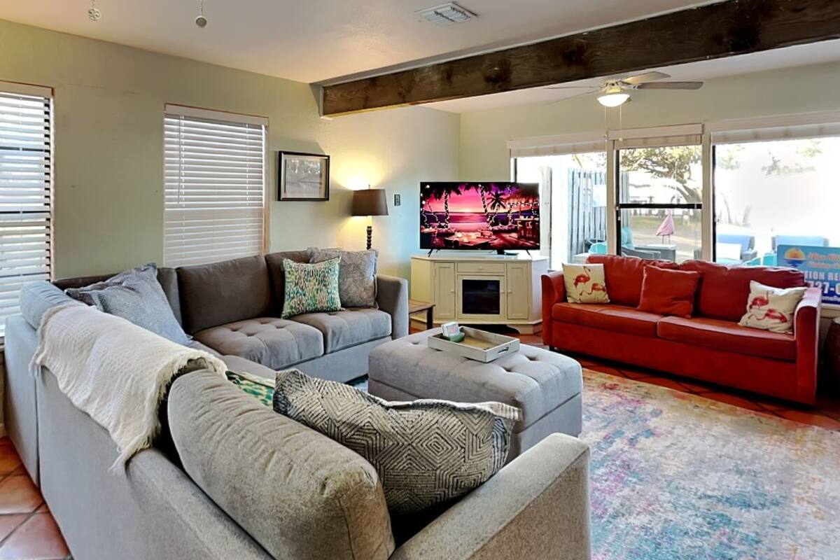 Una sala de estar con un sofá gris, un sofá rojo y un televisor. Hay una ventana con persianas y una lámpara sobre una mesa.