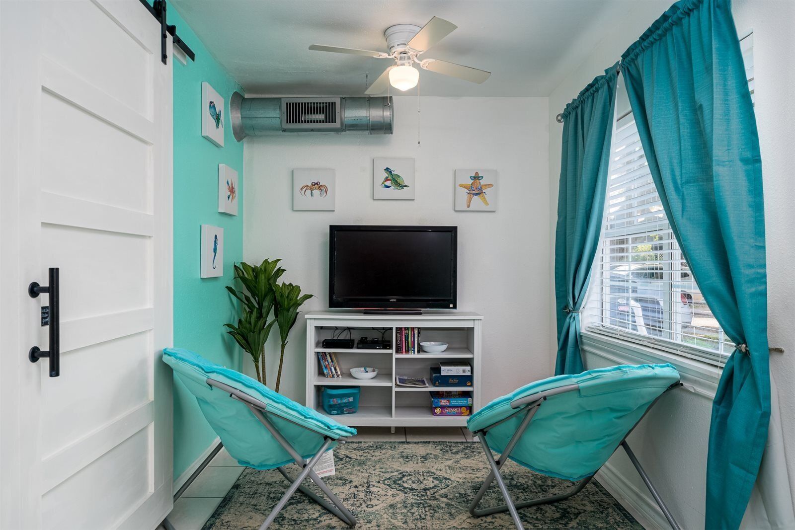 A compact living space with teal walls, a ceiling fan, a flat-screen TV, a white entertainment unit, two teal folding chairs, and a window with teal curtains.