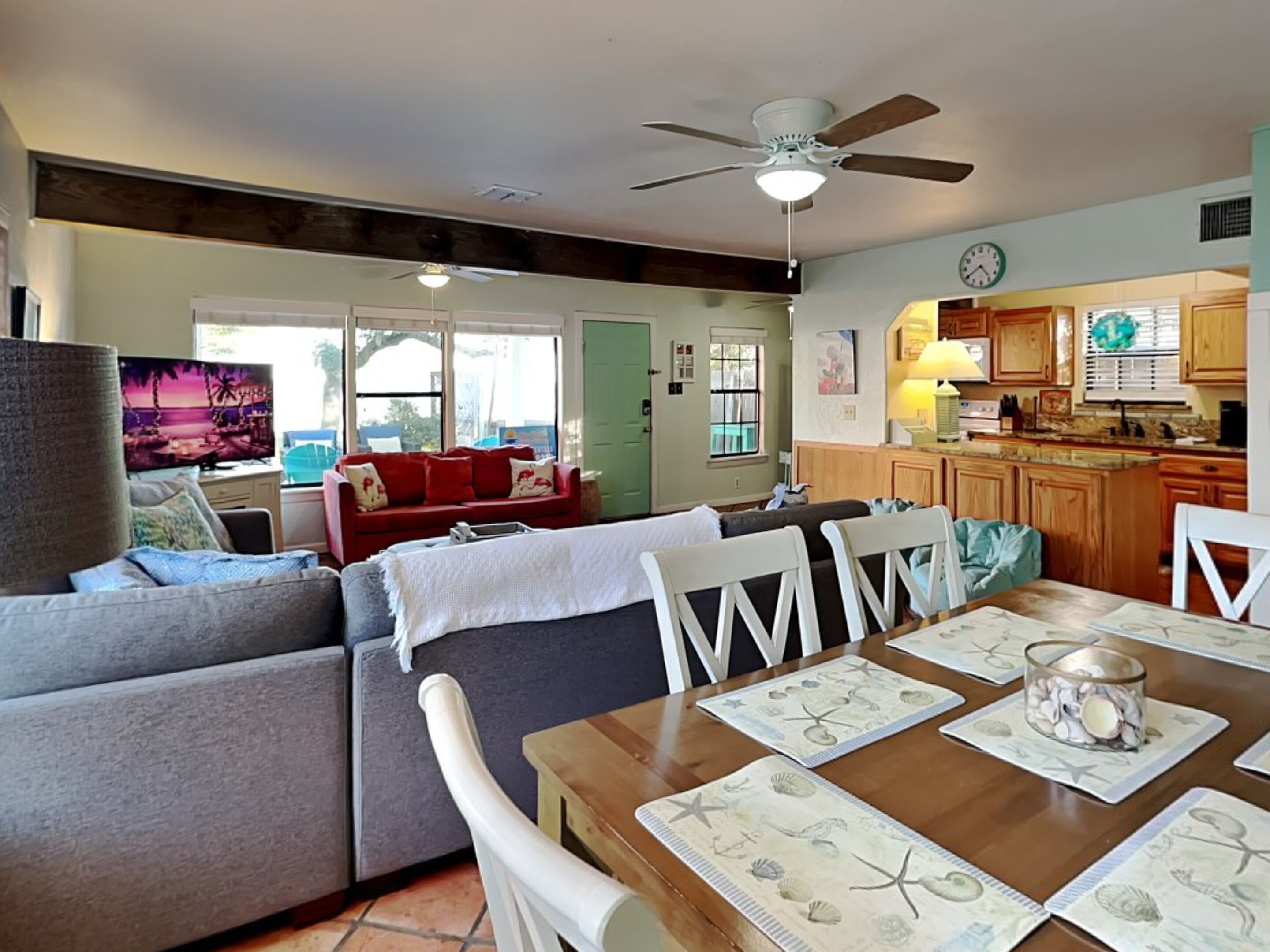 A living room with a sofa, chairs, and a dining table with placemats. There is a kitchen area with wooden cabinets and a clock on the wall.