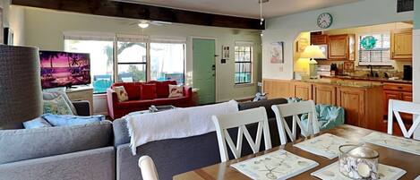 A living room with a sofa, chairs, and a dining table with placemats. There is a kitchen area with wooden cabinets and a clock on the wall.