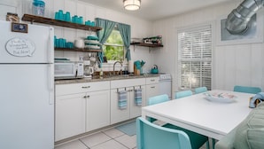 A kitchen with white cabinets, a white refrigerator, a stove, and a dining table with chairs. There are teal-colored items on the shelves and a window with curtains. - Sun Harbor Cottages by Miss Kitty’s (Rockport)