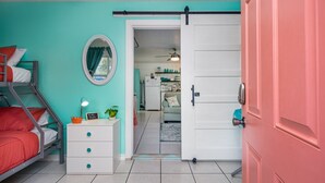A room with bunk beds, a white dresser, a mirror, and a sliding door leading to a kitchen area. - Sun Harbor Cottages by Miss Kitty’s (Rockport)