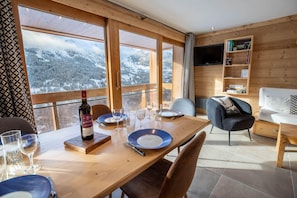 Uma área de refeições com mesa de madeira posta para dois, uma garrafa de vinho e vista para montanhas cobertas de neve através de grandes janelas.