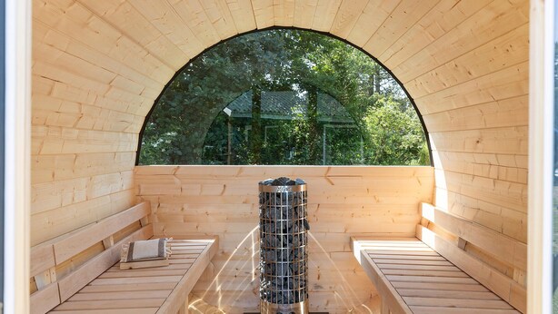 A sauna with wooden benches and a central heater, surrounded by a wooden arch and a large window.