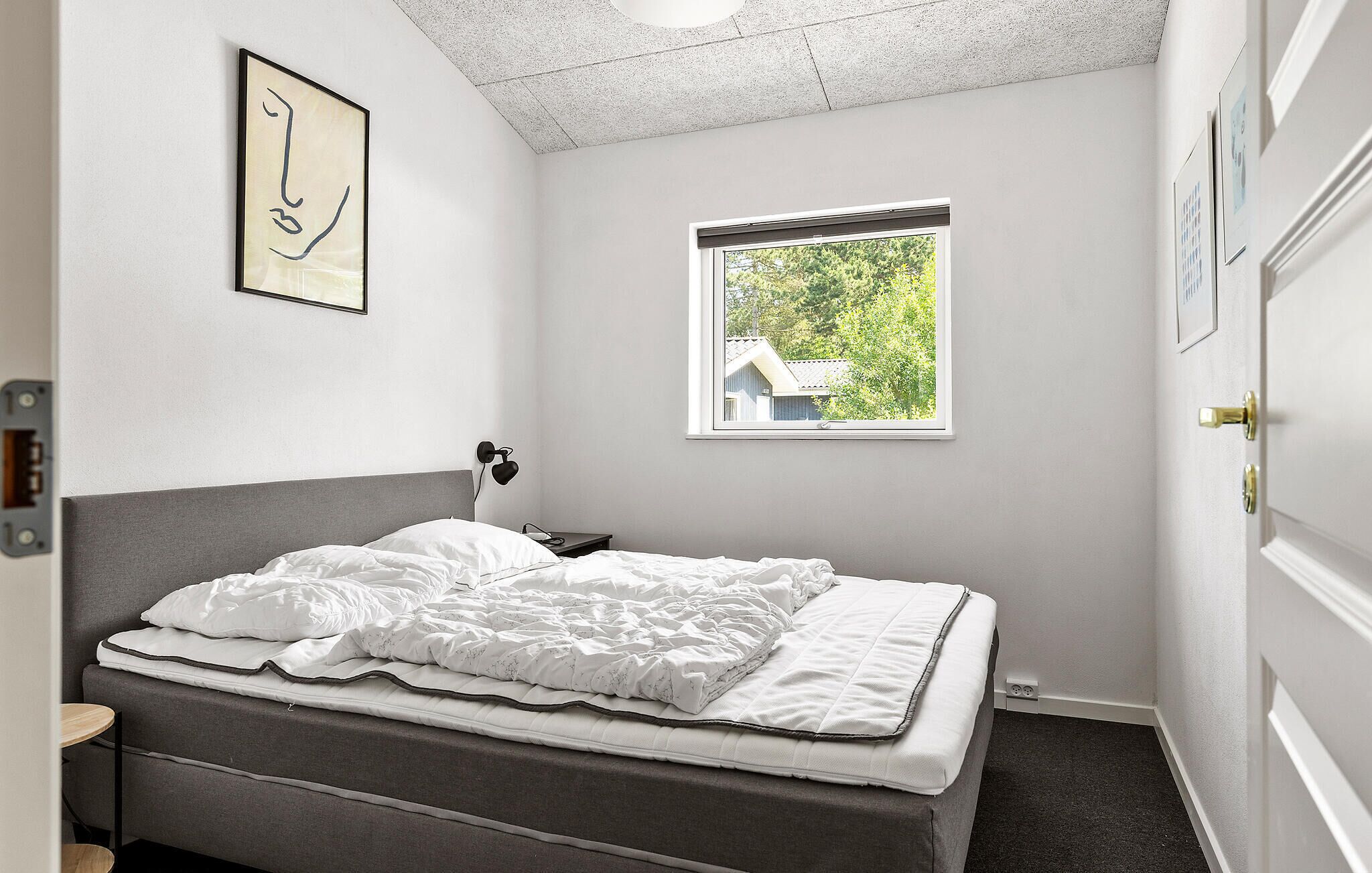Une chambre à coucher comprenant un lit, une table de chevet, une lampe fixée au mur, une fenêtre donnant sur un espace verdoyant et une porte.