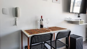 Ein Essbereich mit einem für zwei Personen gedeckten Tisch, einer Weinflasche und zwei Weingläsern. Zwei schwarze Stühle mit perforierten Rückenlehnen stehen bereit. Auf dem Tisch liegt eine gewebte Tischmatte. Eine Telefon und eine Wandleuchte sind an der Wand zu sehen.
