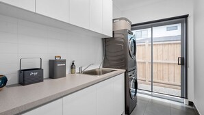 A modern kitchen with white cabinets, a built-in washing machine, and a large sliding glass door leading to a backyard. - Luxury Large Home - LATE CHECK OUT Feb-Nov 2025 (Point Lonsdale)