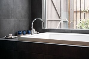Une salle de bain moderne avec un grand lavabo blanc, des murs recouverts de carreaux foncés et une fenêtre donnant sur une clôture en bois et de la verdure.