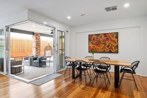 Un espace repas moderne avec une table en bois et des chaises noires, une porte vitrée donnant sur un coin salon extérieur, et une peinture abstraite aux couleurs vives accrochée au mur.