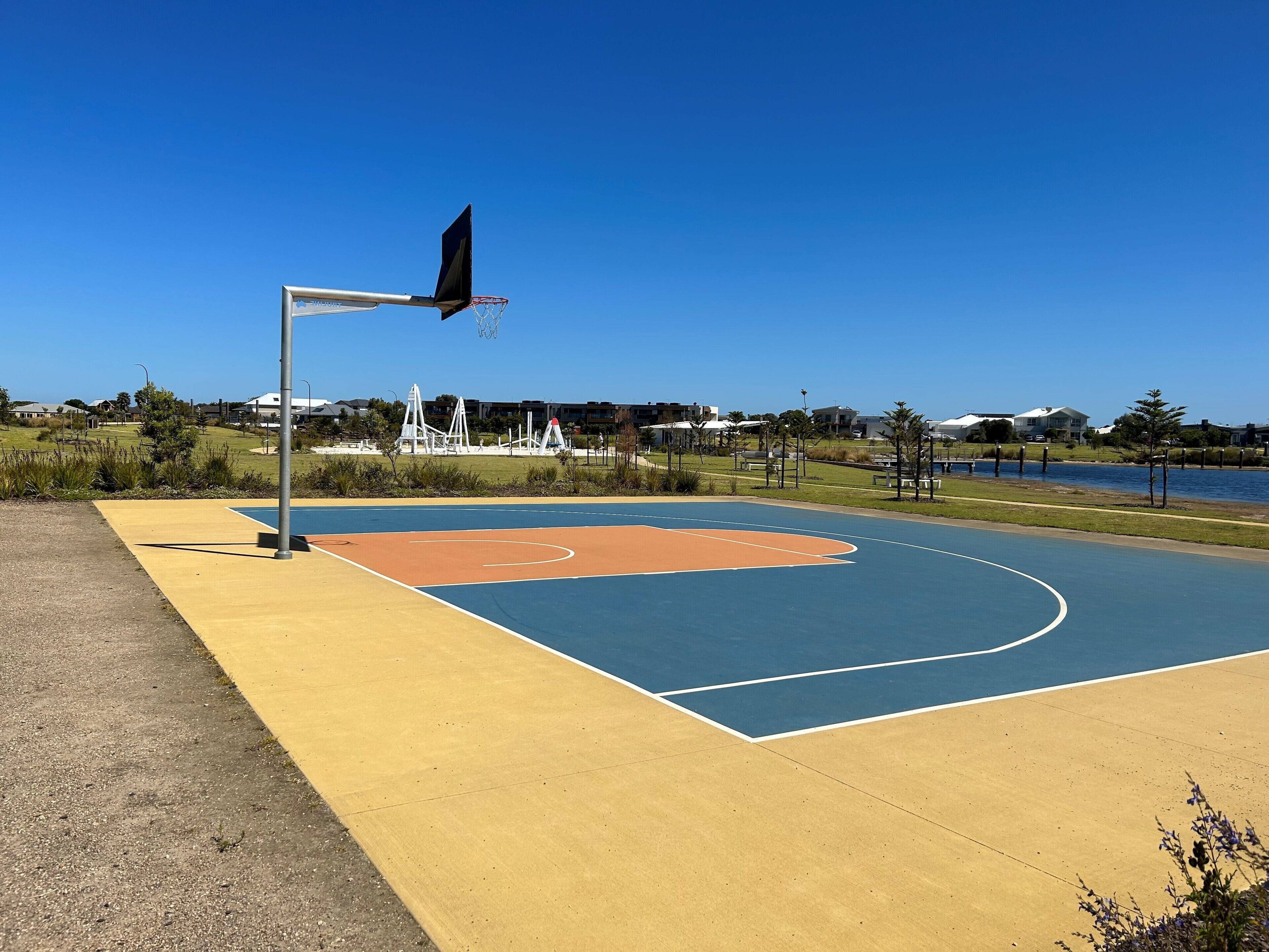 An outdoor basketball court with a blue and orange playing surface, surrounded by grass and trees.