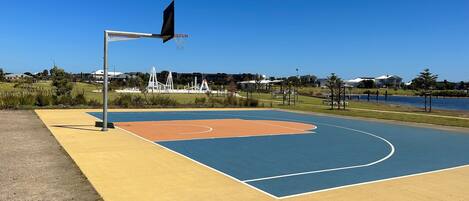 An outdoor basketball court with a blue and orange playing surface, surrounded by grass and trees.