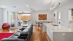 A modern living space with a black leather sofa, a red chair, a dining area with a table and chairs, and a kitchen with a white countertop and sink. - Luxury Large Home - LATE CHECK OUT Feb-Nov 2025 (Point Lonsdale)