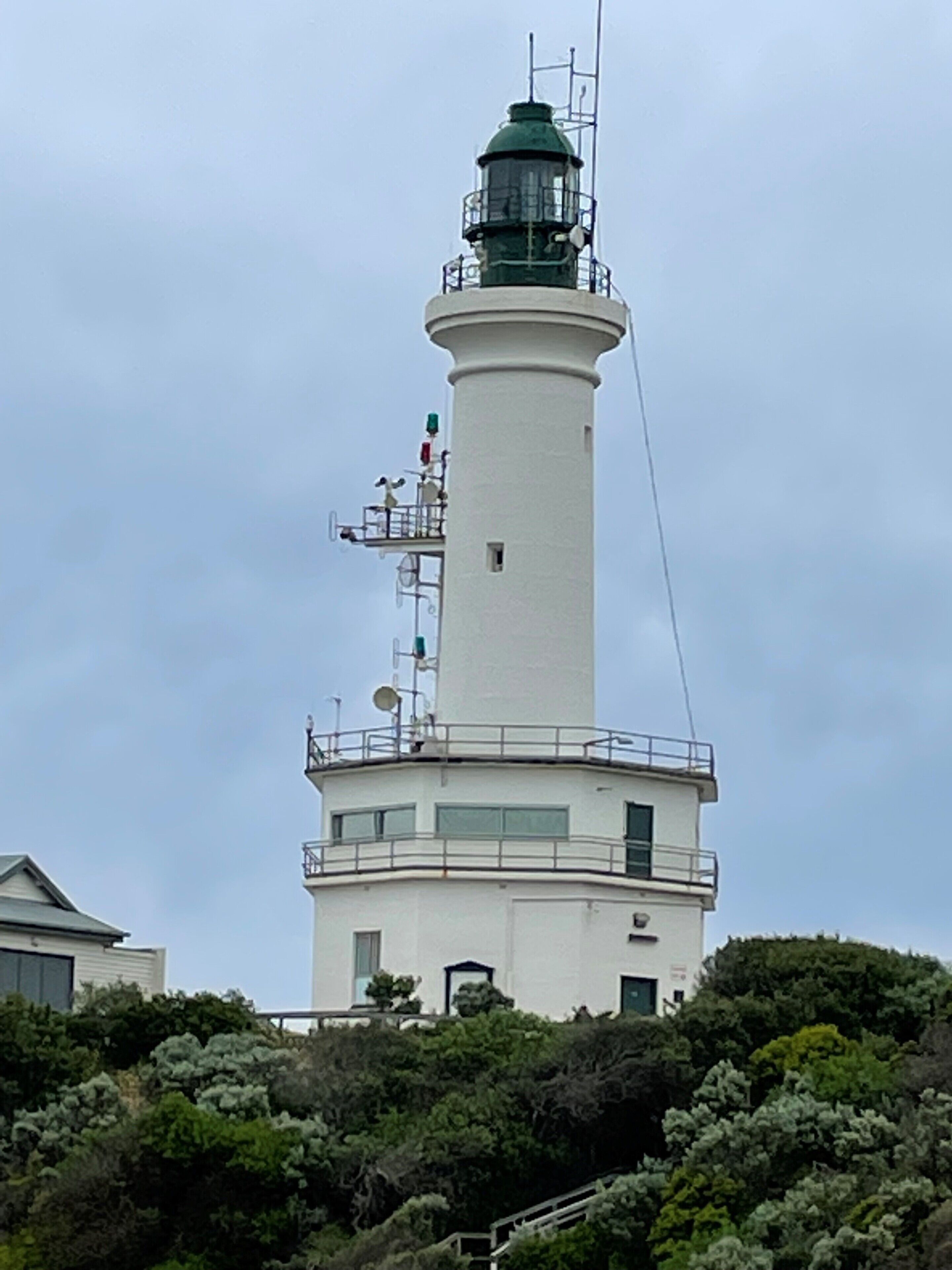 Um farol com torre branca e topo verde, rodeado por vegetação densa.