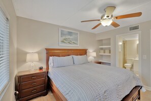 A bedroom with a wooden bed, nightstands, a ceiling fan, and a painting on the wall. - St Augustine Beach Condo just steps from the beach! (St. Augustine)