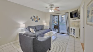 A living room with a grey sofa, a white coffee table, a ceiling fan, a TV on a cabinet, and a view of the outside through a glass door. - St Augustine Beach Condo just steps from the beach! (St. Augustine)