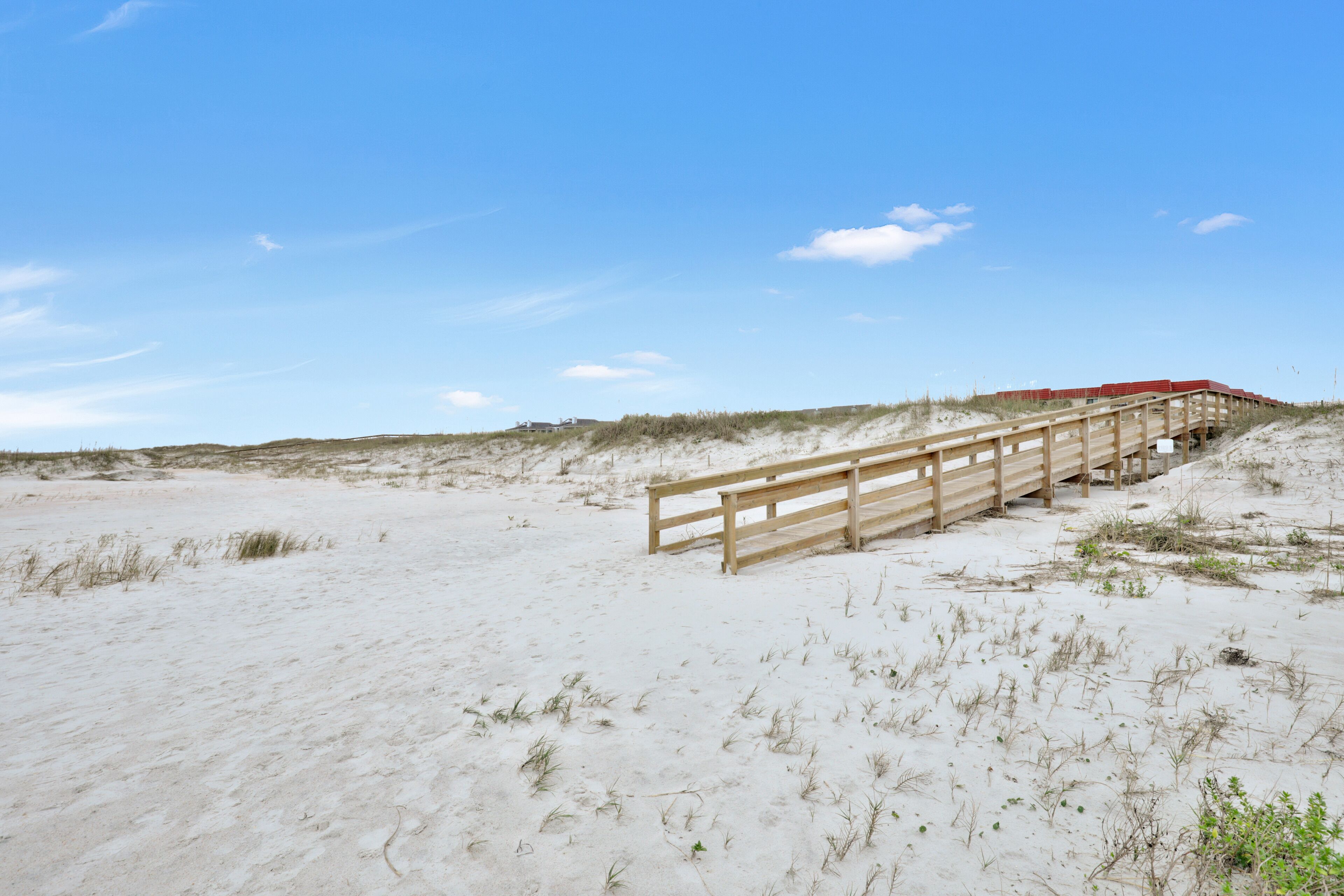 Un muelle de madera que conduce a una estructura de techo rojo sobre una playa arenosa, con dunas al fondo.