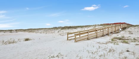 Un muelle de madera que conduce a una estructura de techo rojo sobre una playa arenosa, con dunas al fondo.