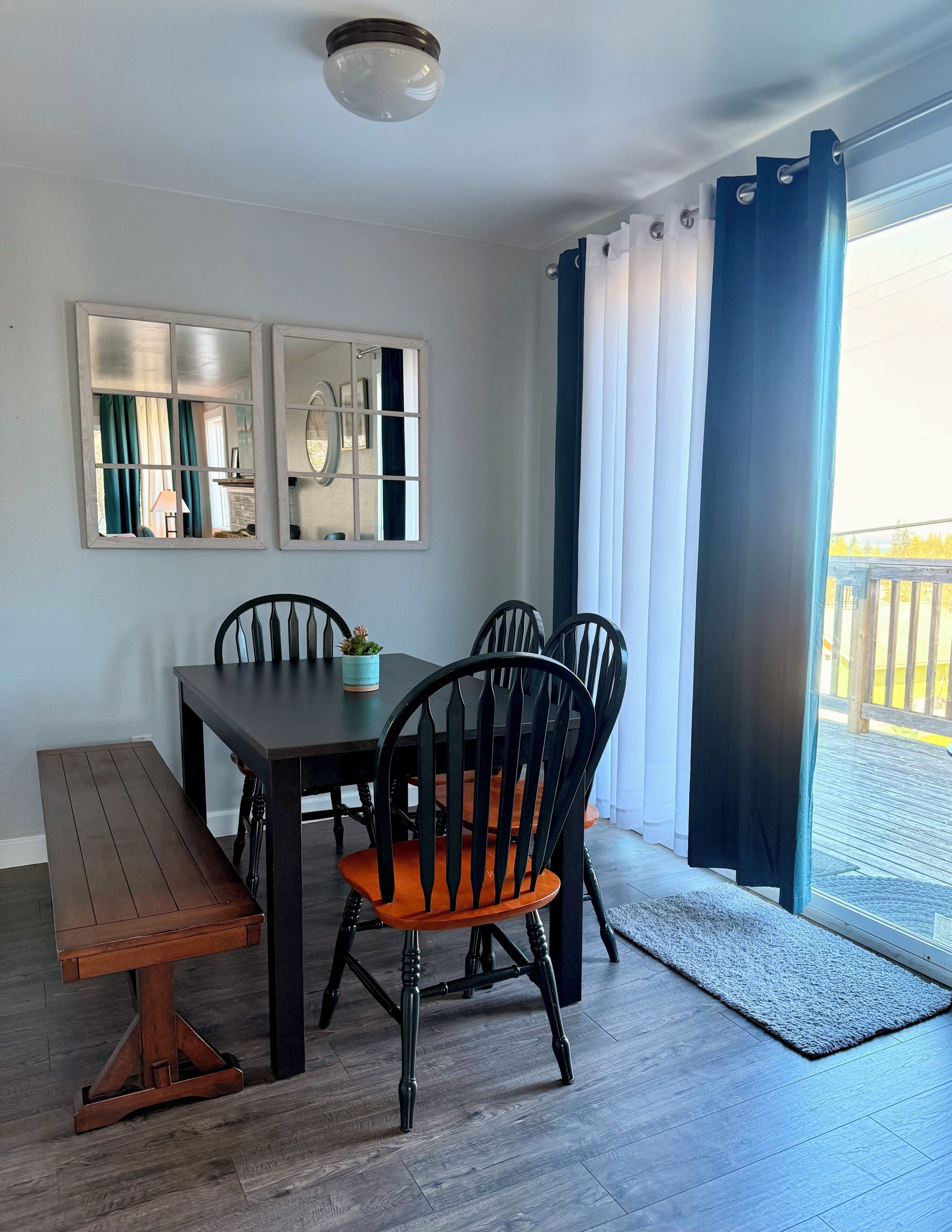 A dining area with a wooden table, four chairs, a bench, two wall-mounted mirrors, and a view through a large window with curtains.