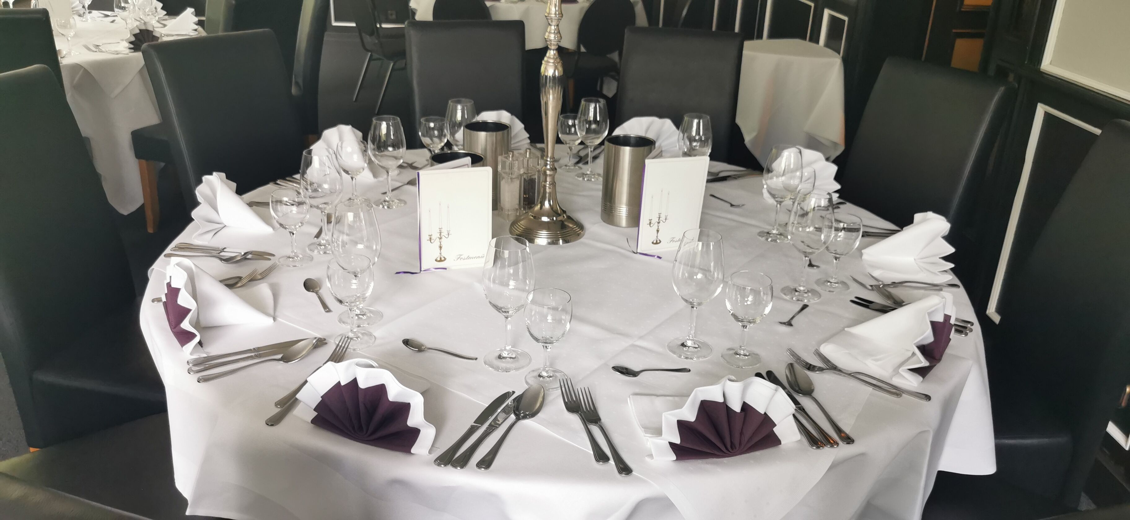 A dining table set for a formal meal with multiple wine glasses, cutlery, and a menu stand.