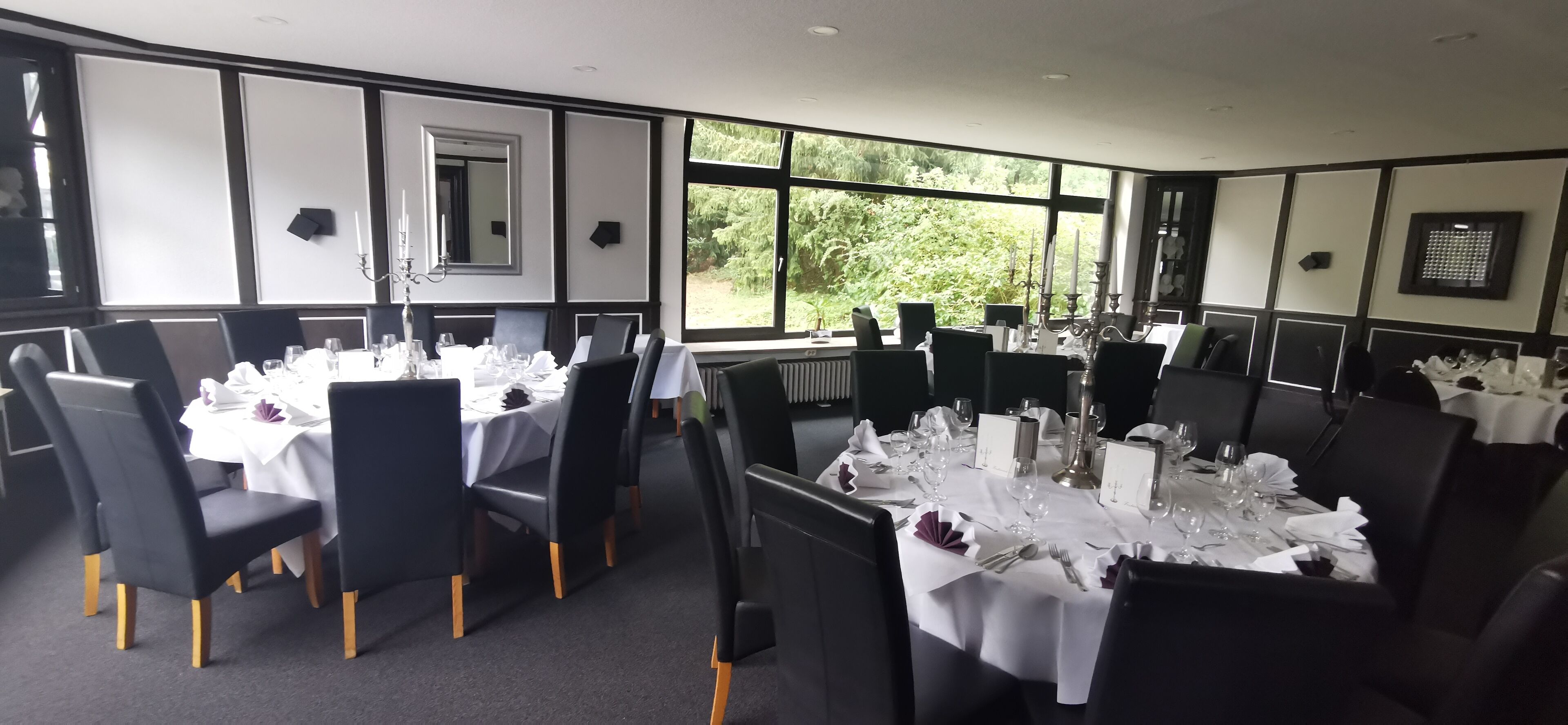 Un espace repas avec plusieurs tables dressées pour un repas, comprenant des chaises noires, des nappes blanches et des chandeliers.