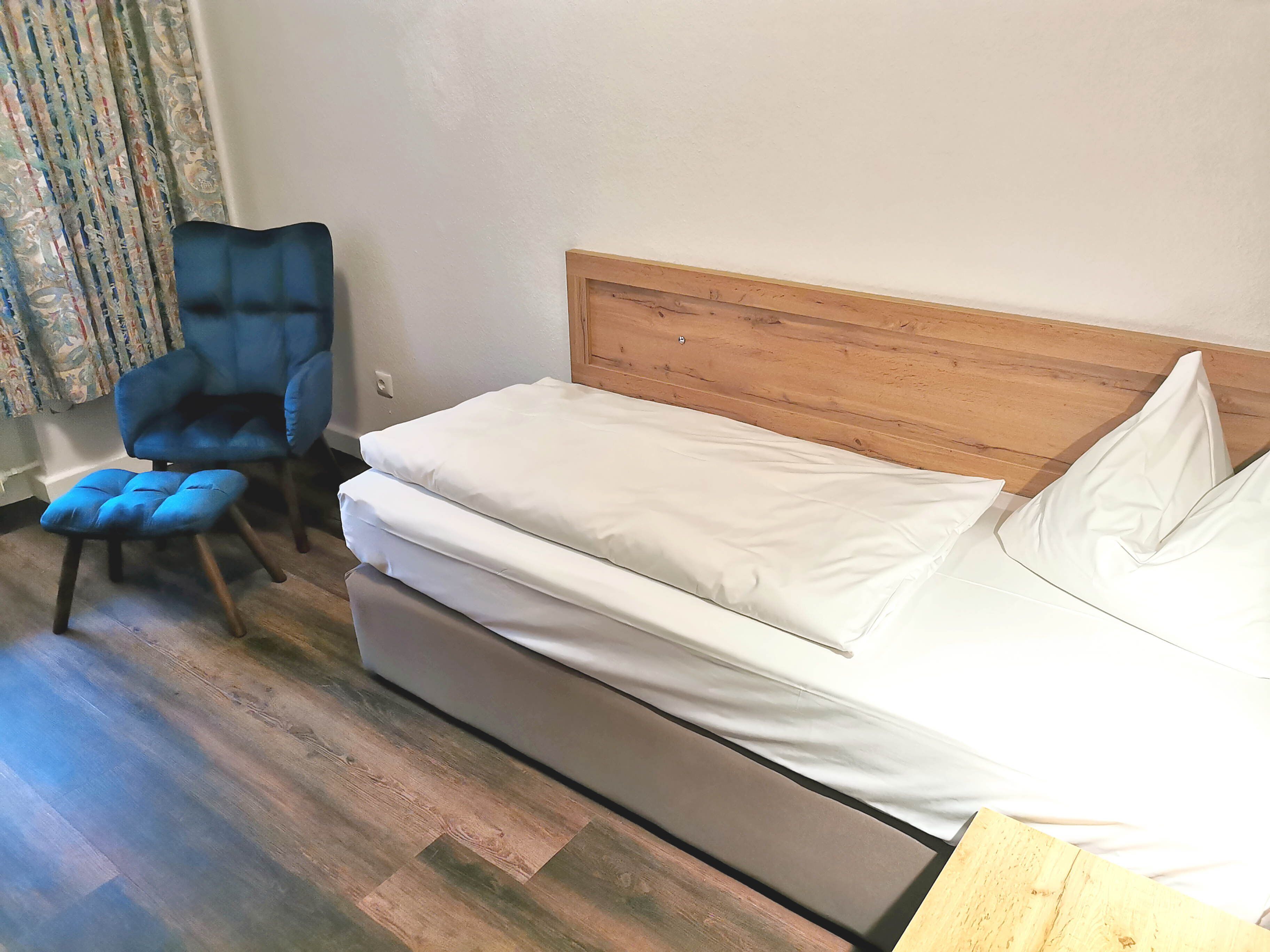 A single bed with a wooden headboard, a blue chair, and a patterned curtain.